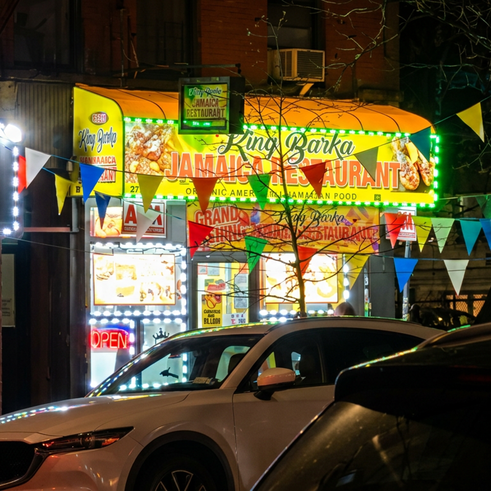 Nighttime view of King Barka Jamaican restaurant storefront decorated with colorful pennant flags and bright illuminated signs.