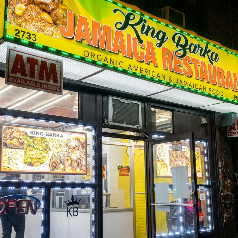 Nighttime storefront of King Barka Jamaica Restaurant, featuring brightly lit signage, food images, ATM notices, and an open sign.